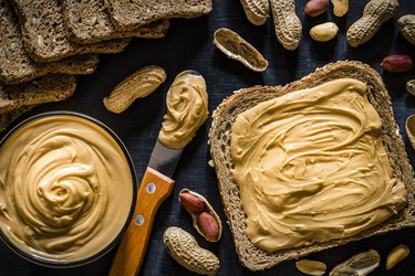 Tryptophan-rich peanut butter spread on a slice of bread with a knife of peanut butter and peanuts on a dark surface
