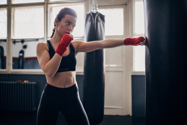 a person wearing black leggings, a black sports bra and red boxing wraps punching a bag at the gym