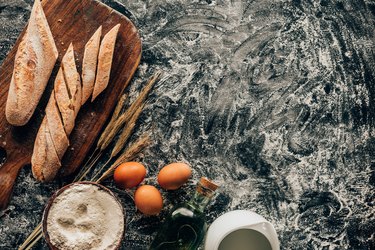 top view of arranged pieces of french baguette, raw eggs, wheat and flour in bowl on dark surface