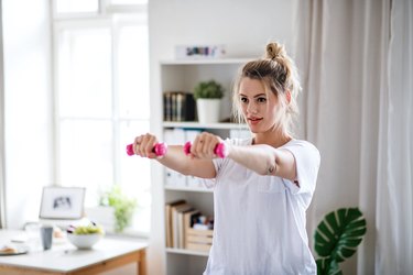Young woman with dumbbells doing exercise in bedroom indoors at home