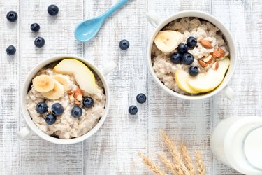 Two white bowls of oatmeal topped with sliced almonds apples and blueberries on a white table