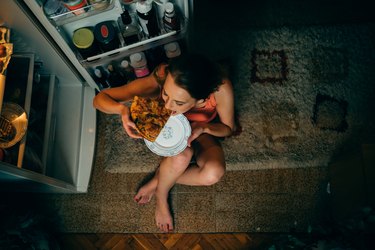 Woman eating in front of the refrigerator in the kitchen late night, as a reason for waking up bloated