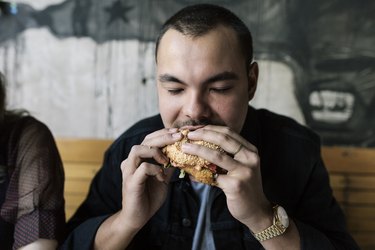 a person wearing a black button down shirt and a blue tie eats a burger sitting on a wooden bench