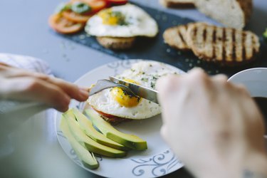 a close up of a person's hands cutting into a breakfast of eggs, avocado slices, bread and tomatoes
