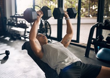 person with short hair and tattoos on their biceps demonstrates how to do the dumbbell bench press at the gym