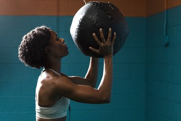 Side view of woman holding a slam ball while listening music against wall in gym