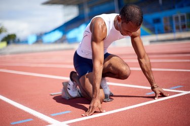 an athlete wearing a white sleeveless shirt and shorts crouches on a track about to run in hot sun