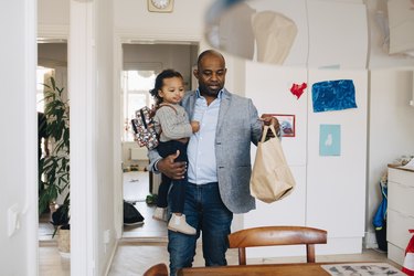 Father carrying daughter with backpack while keeping paper bag on table at home