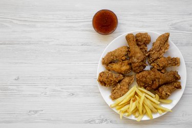 Tasty fried chicken legs, spicy wings, French fries, chicken tenders and sauce on white plate over white wooden background, top view. Flat lay, overhead, from above. Copy space.