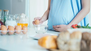 close up of pregnant person in blue dress cracking an egg into a bowl standing in kitchen making gestational diabetes breakfast
