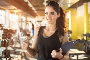 Personal trainer with clipboard showing thumb up in gym.