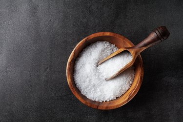 Salt in wooden bowl on black kitchen table top view.