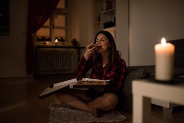 Hungry woman having a slice of pizza at night on couch with candle lit may get the cookies sweats or feel sweaty after candy