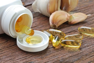 Garlic oil capsules spilling out of a white pill bottle on a wooden table with garlic cloves in the background