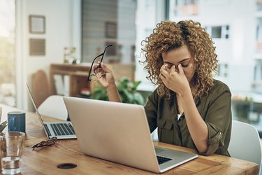 woman with headache holding eyes and glasses at table with two laptops