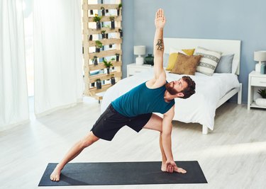 man with beard wearing blue tank top practicing yoga on a black mat in his bedroom