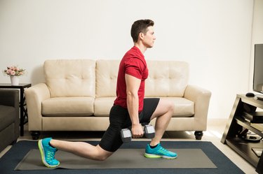 fit man wearing a red shirt and black shorts and doing a reverse lunge with dumbbells in living room