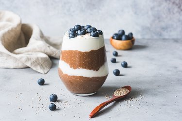 A glass of Chocolate Chia Seed Pudding with blueberries on top on a marble counter next to a red spoon and white dish towel