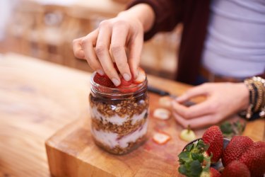 a close up of a person's hands doing breakfast meal prep with yogurt granola and fruit in a mason jar on a wooden cutting board on a counter