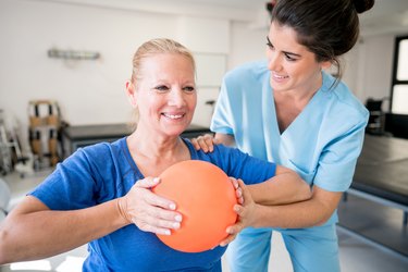 Senior woman patient at physical recovery therapy exercising with a ball and therapist helping her