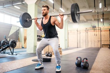 an athlete wearing a black tank top and gray sweat pants does a barbell exercise in the gym in front of two kettlebells