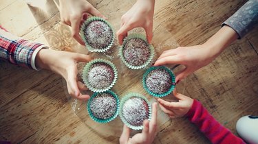 an overhead photo of six people's hands taking cupcakes off a wooden table
