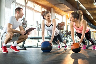 Two sporty girls exercising with fitness balls while their fitness instructor tracking the progress on clipboard.