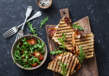 Grilled bacon, mozzarella sandwiches on wooden cutting boards and arugula, cherry tomato salad on dark background, top view.Delicious breakfast or snack, flat lay