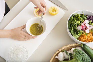 overhead photo of a person's hands as they whisk a fresh salad dressing with lemon in a small white bowl next to a bowl of salad