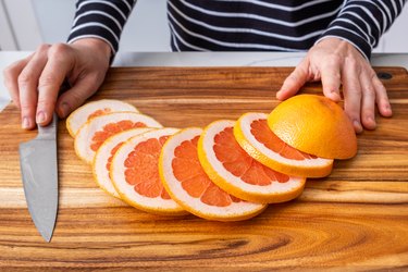 Caucasian woman hands placed on wooden chopping board with sliced red grapefruit and knife