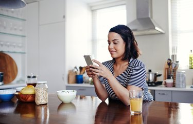 Woman on phone tracking net carbs in the kitchen surrounded by orange juice and oatmeal