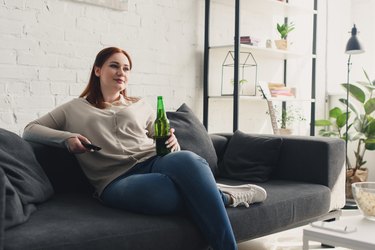 beautiful woman holding bottle of beer and remote control at home