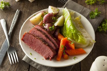 an overhead photo of a white plate of homemade roaster oven corned beef with cabbage, carrots and potatoes on a wooden table next to a silver fork and knife