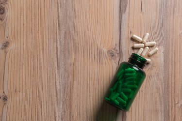 white capsules coming out of green supplement bottle on wooden background