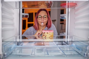 Woman eating gateau while standing in front of fridge.