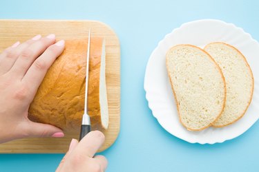 Woman's hands cutting loaf of white bread with knife. Slices on plate on pastel blue desk. Preparing breakfast. Point of view shot. Closeup. Top view.