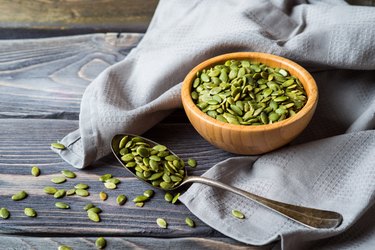 A wooden bowl filled with pumpkin seeds surrounded by a grey cloth, with a spoonful of pumpkin seeds on top of a grey wooden table.