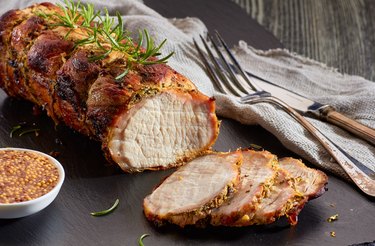 Roasted pork loin with whole grain mustard on a cutting board next to knife and fork.