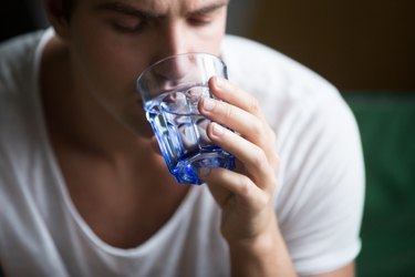 a photo of a person wearing a white t-shirt feeling thirsty and drinking water from a small holding blue glass