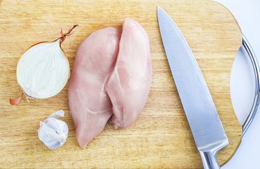 Top View of Raw Chicken Breasts, Fillets on a Wooden Cutting Board with Ingredients, Chopped Half Onion, Whole Garlic Bulb, and Stainless Chef Knife. Organic Food, Poultry Meat Cooking Concept