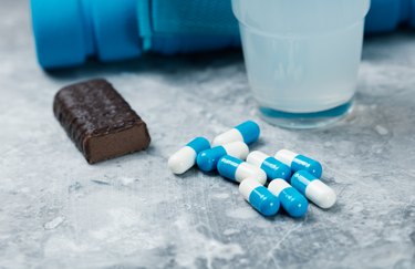 Creatine capsules, High Protein Bar, BCAA (amino acids) Drink and blue dumbbell in background. Sport nutrition. Stone background. Close up. Copy space.