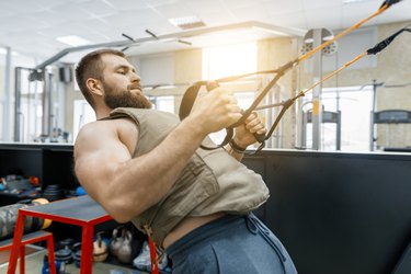 Muscular bearded man dressed in military weighted armored vest doing exercises using straps systems in the gym. Sport, training, bodybuilding and healthy lifestyle concept.