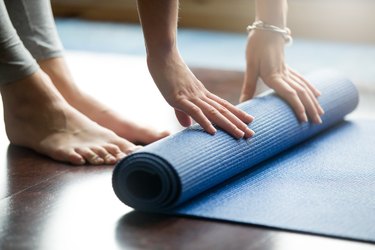 woman rolling up blue yoga mat on wooden floor with bare feet