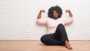 Young african american woman sitting on the floor at home showing arms muscles smiling proud. Fitness concept.