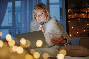 an adult with short blond hair holds a glass bowl of a healthy late night snack while watching a show on their laptop with string lights in the background