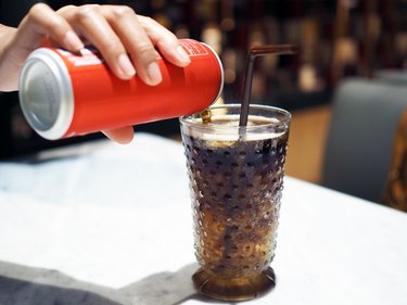 Pouring cola over ice cube in clear glass with straw.