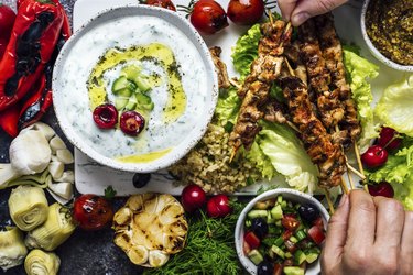 Hand taking chicken skewers from a board with lots of snacks and a bowl of yogurt and cucumber dip for savory yogurt recipes.