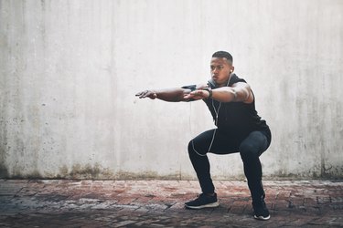man wearing black workout clothes and headphones doing body-weight squat outside in front of gray wall