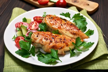 Two pieces of baked chicken breast on a plate with herbs and tomatoes