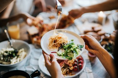 Group of joyful young man and woman having fun, passing and sharing food like carbs across table during party